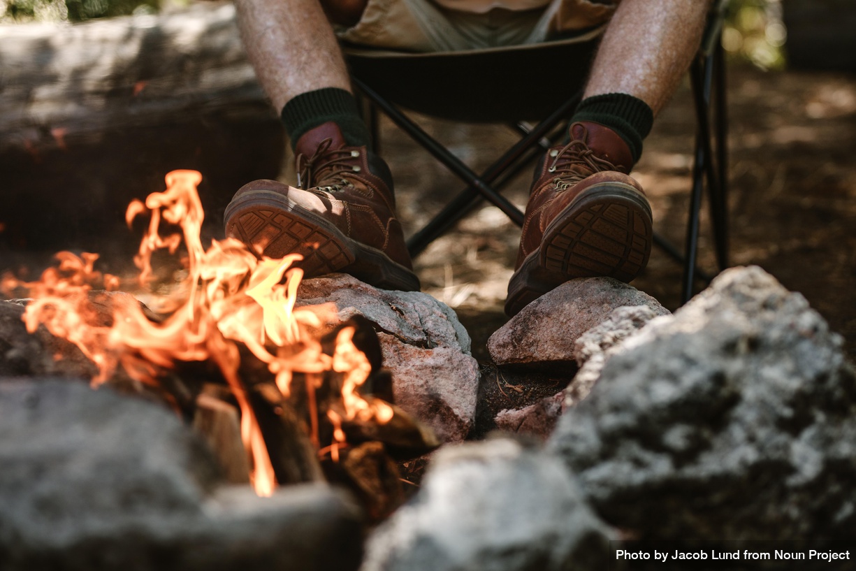 Older man in boots warms his feet by the fire at camping site by Jacob Lund from Noun Project
